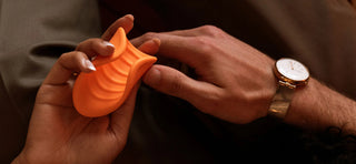 Close up of couple's hands, one person holding orange vibrating ribbed stroker, other person wearing watch and holding other person's hand