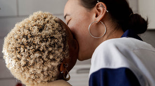 Close up of two women kissing, one with short bleached curly hair and the other with black hair in a bun
